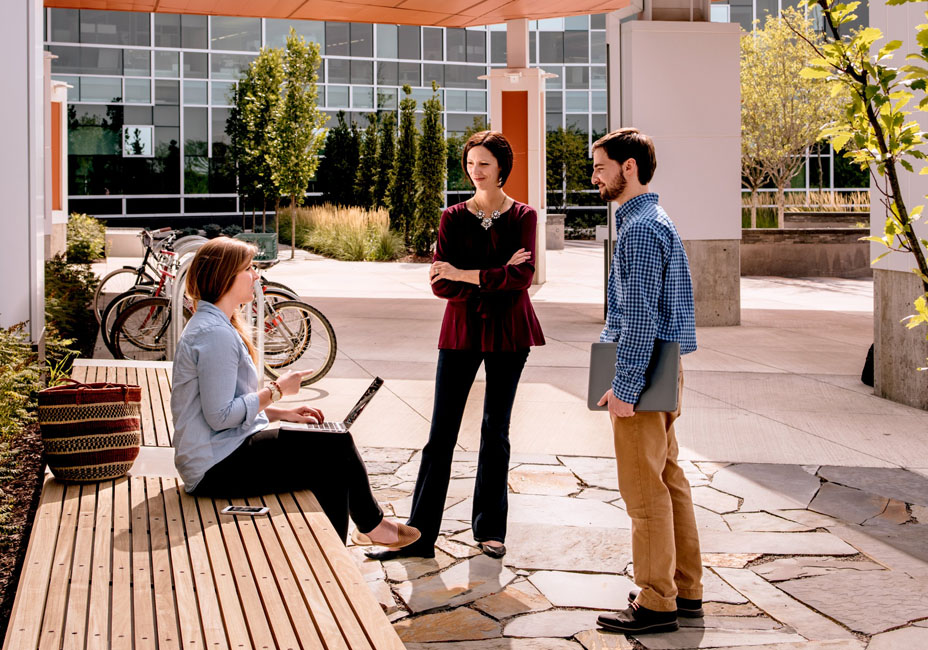 One male and one female talking to a women sitting with a laptop on her lap outside of a building. 