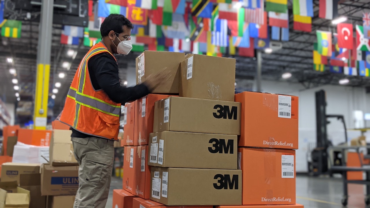 Direct Relief employee wearing a safety vest and mask stacks boxes of 3M product in a warehouse.