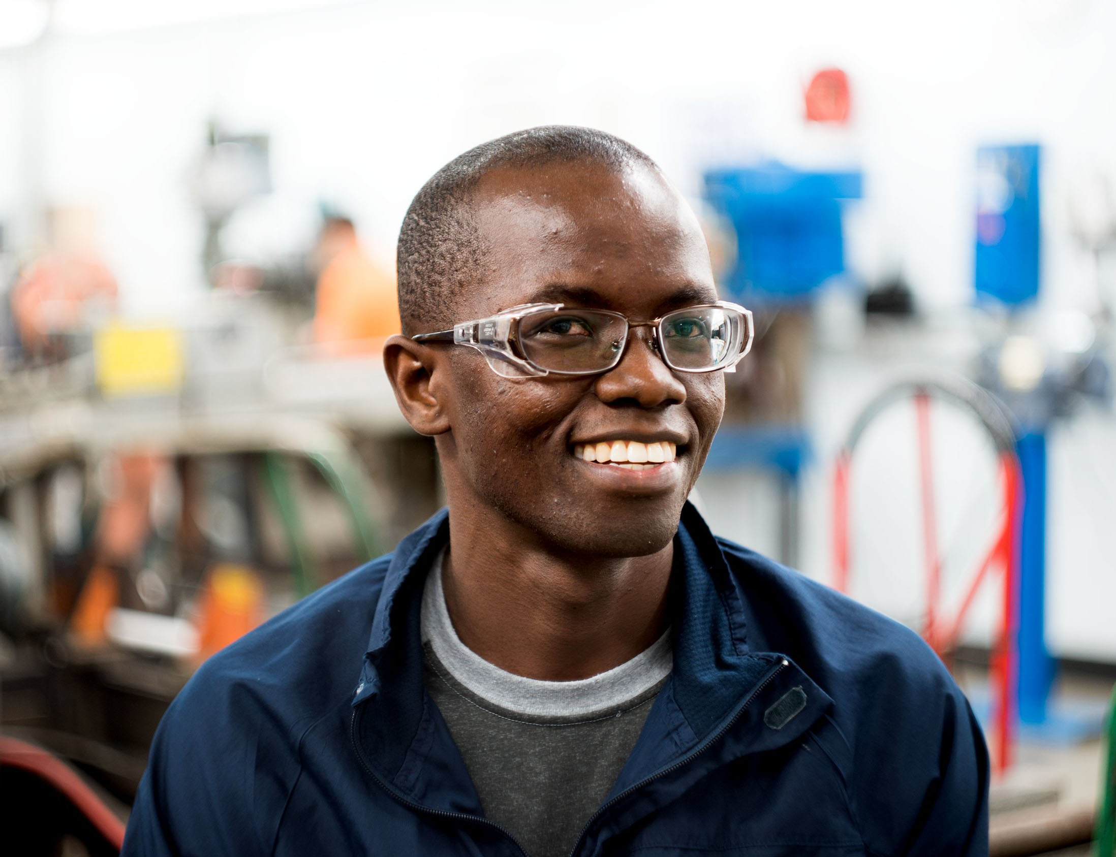 Young man smiles while wearing safety glasses. Manufacturing equipment blurred in the background.