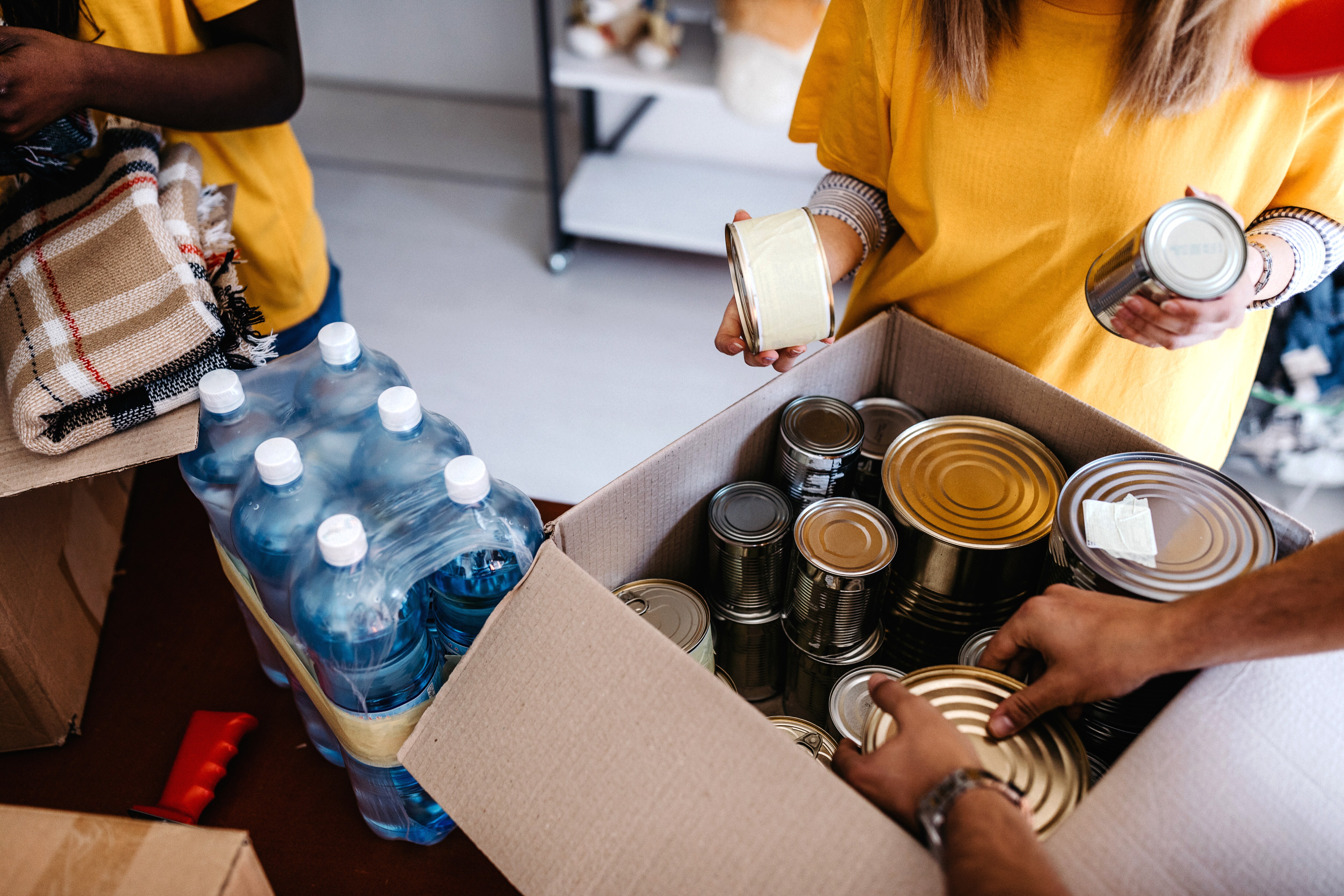 People pack canned goods into a box. A case of water sits next to the box on the table.