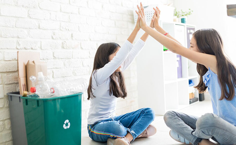 Two girls hold hands above their heads with a recycling bin behind them.